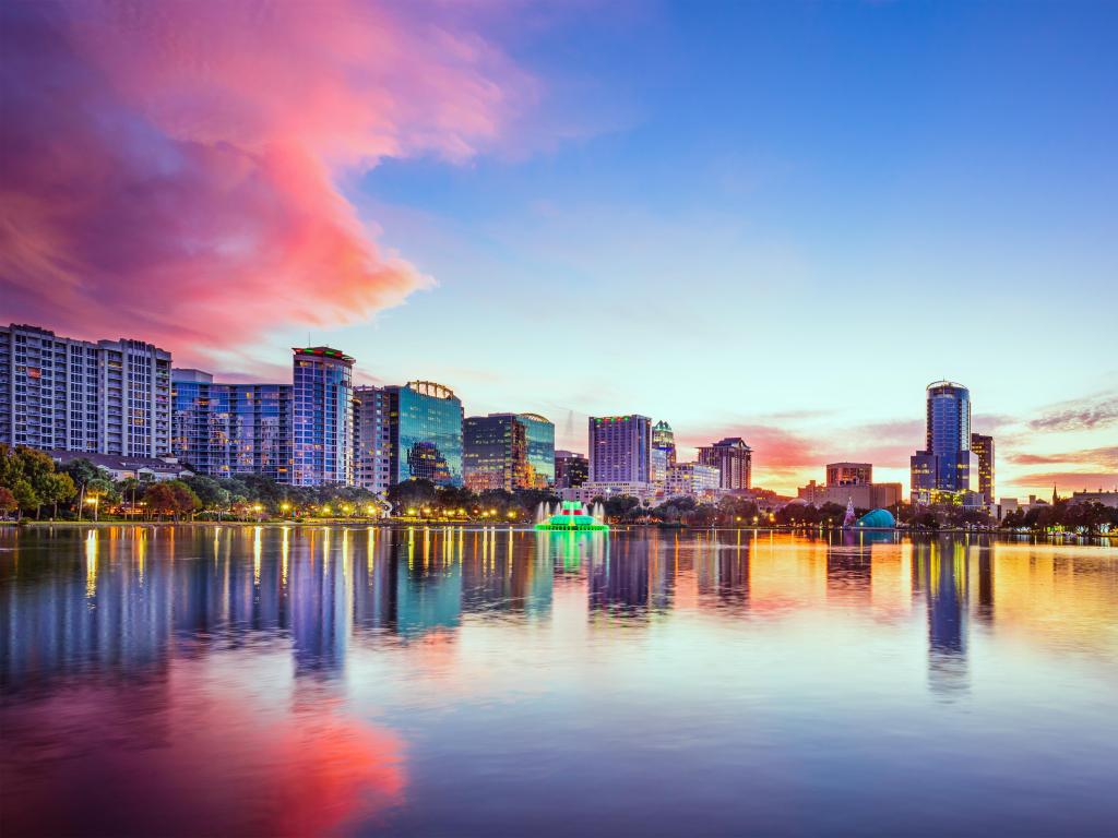 Orlando, Florida, USA downtown city skyline on Eola Lake at sunset with the city buildings reflecting in the water and a pink hue in the sky.