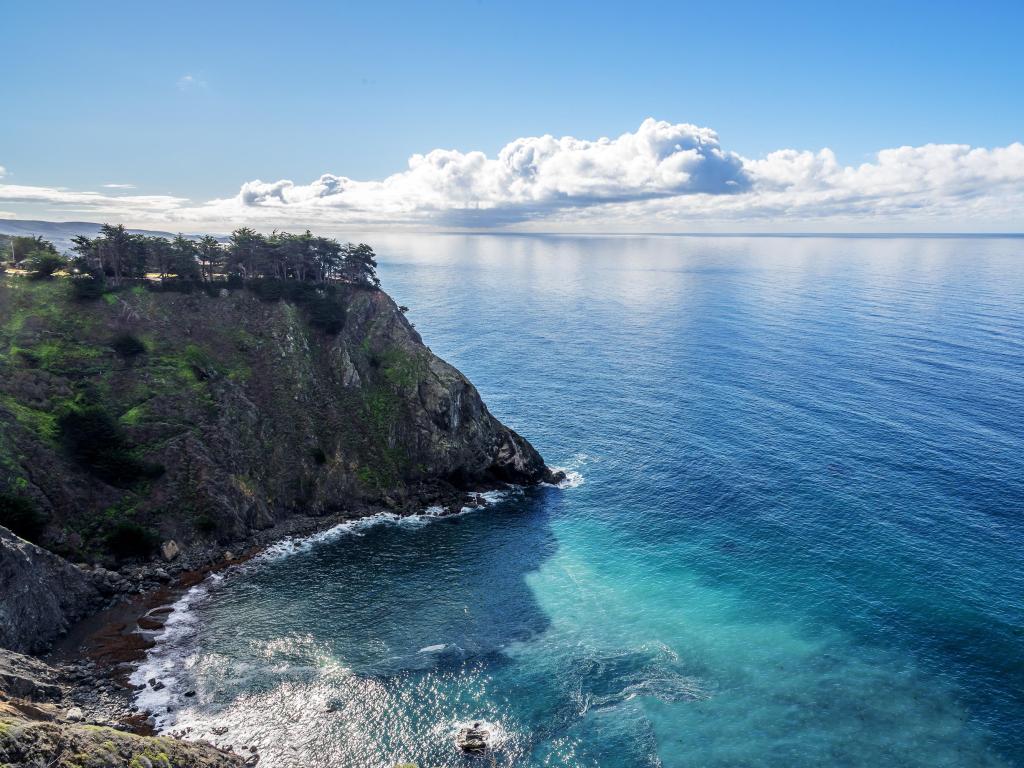 Shimmering reflective sunlight, blue skies, white clouds and waves splashing along the ragged cliffs of Ragged Point by Highway 1 on the California Central Coast.