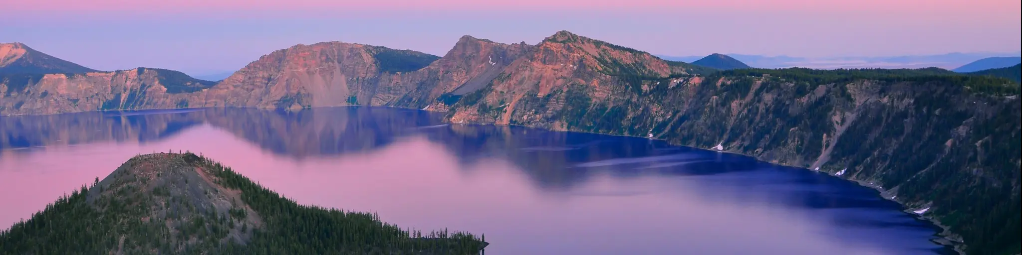 Oregon, USA with a beautiful view of Crater Lake at sunset with hills in the distance and a pink sky.
