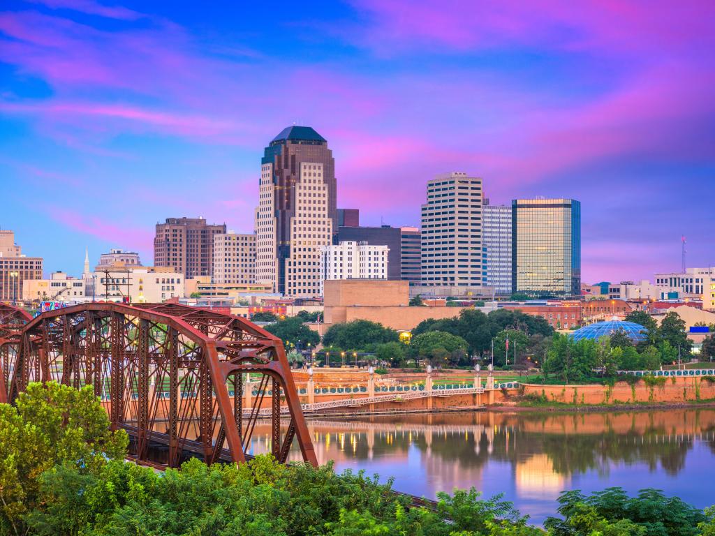 Shreveport, Louisiana showing the downtown skyline in the distance, and a metal bridge crossing the Red River in the foreground at sunset.