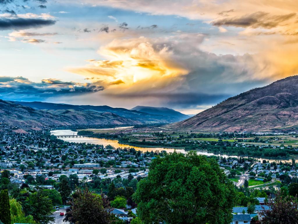 Kamloops, Canada with the pretty city in the foreground, a river running alongside it and the mountains in the distance with a dramatic sky at sunset. 