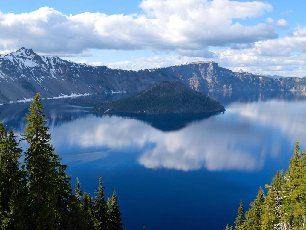 Crater Lake National Park, Oregon, USA taken on a sunny day with the mountains in the distance and lake and trees in the foreground. 