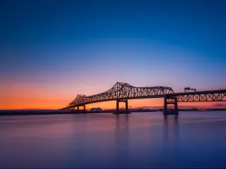 Long bridge over wide river shown in silhouette in blue and orange sunset light