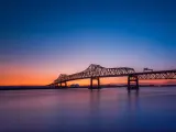Long bridge over wide river shown in silhouette in blue and orange sunset light
