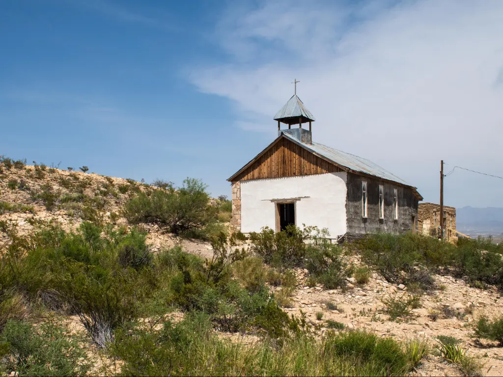 Small white church on dry dirt hillside with bushes and cloudy blue sky