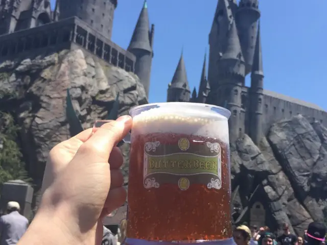A hand raising a cup of Butterbeer up with the replica of the Hogwarts Castle in the background at Universal Studios LA