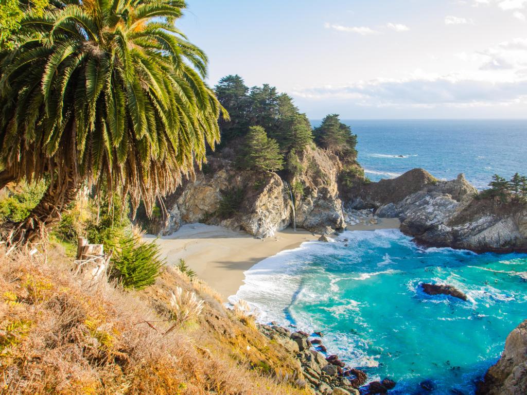 Big Sur, California taken from a hill looking down at a secret waterfall and sand beach surrounded by rocks on a sunny day.