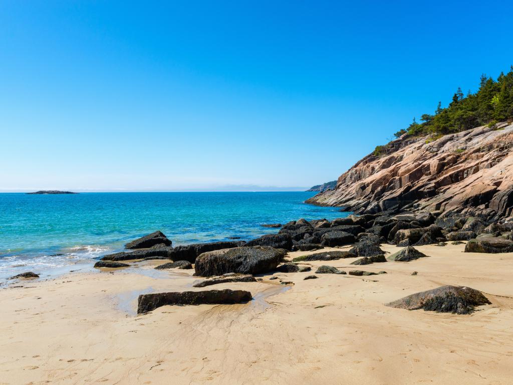 Sand Beach in Acadia National Park, Maine on a sunny day with sand and rock formations to the right and a blue sea in the background. 