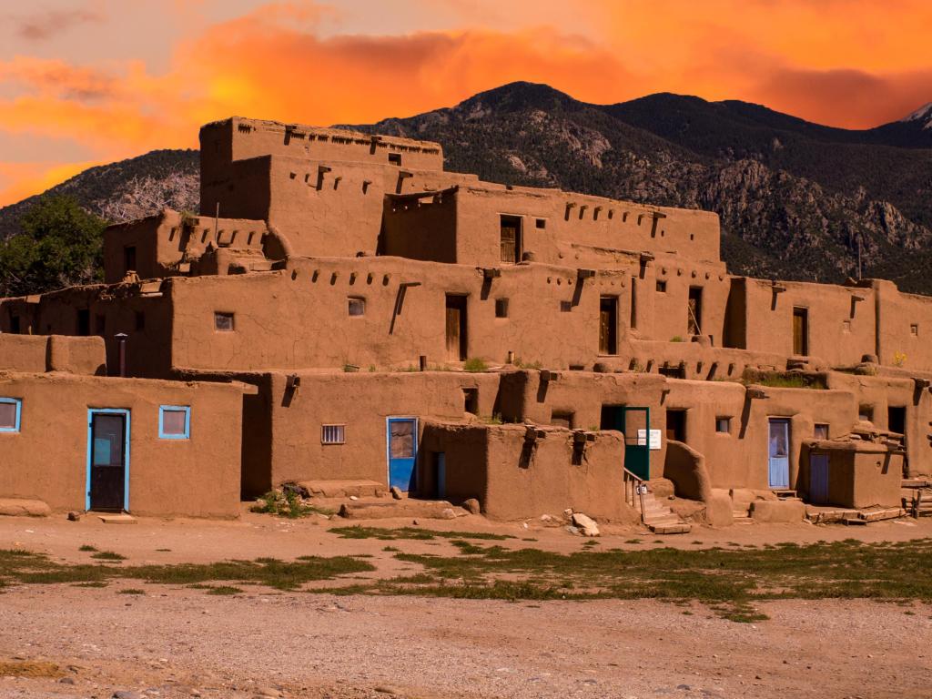 Ancient houses sit atop a hill in Taos, New Mexico, with a vivid orange sunset sky behind