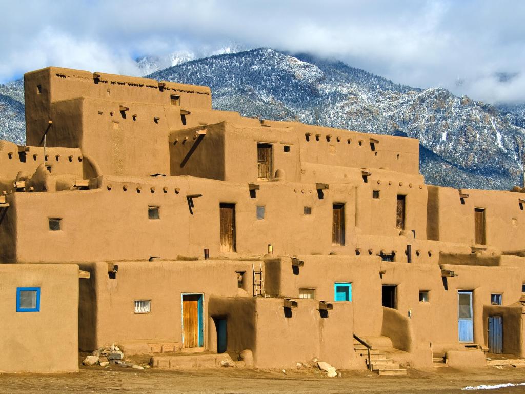 Taos Pueblo in New Mexico, USA with snow-capped mountains in the distance and the mud dwellings in the foreground.