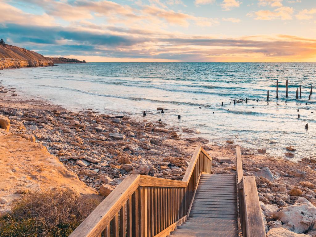 Port Willunga, South Australia with a beach view with a wooden jetty leading to the rocky beach at sunset.