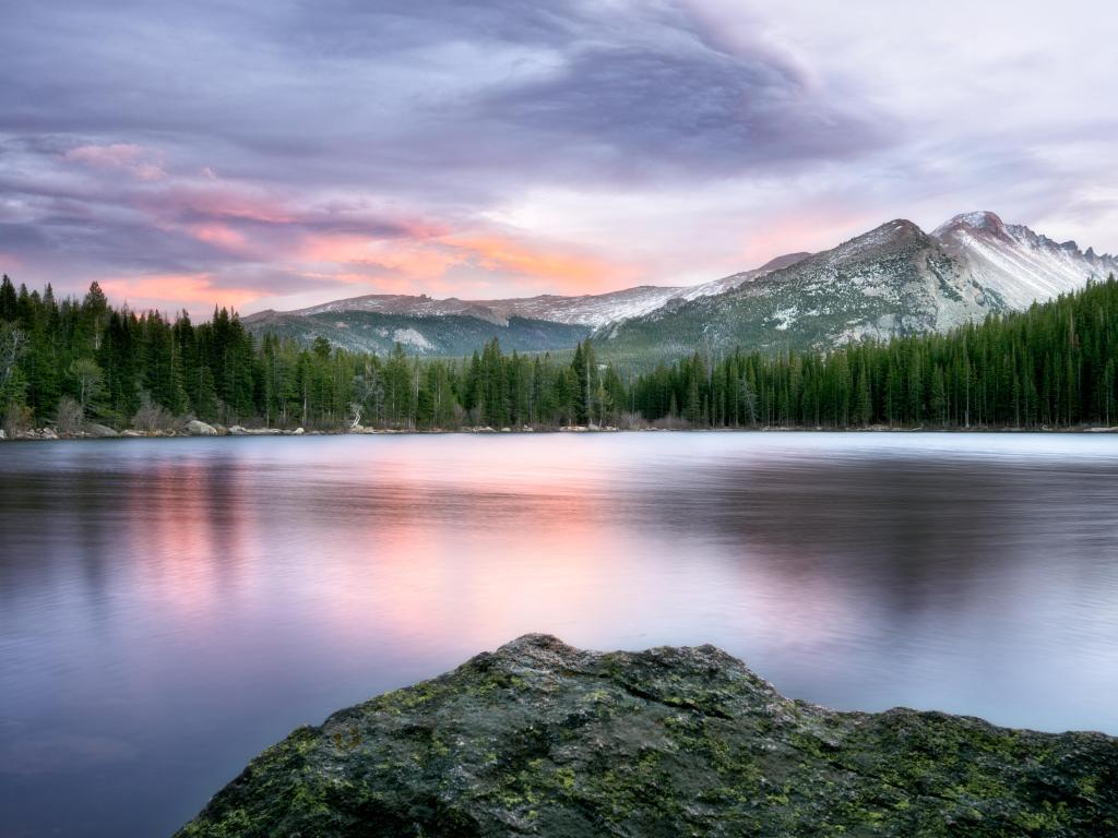 Flat grey water on wide lake reflecting pink sunset light with snow covered mountain behind and pine trees on the lake shores