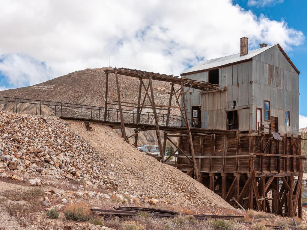 Tonopah Historic Mining Park. Dark wooden and gray corrugated metal sheet building above mine pit under blue cloudscape. Beige stripping or waste rock heap.