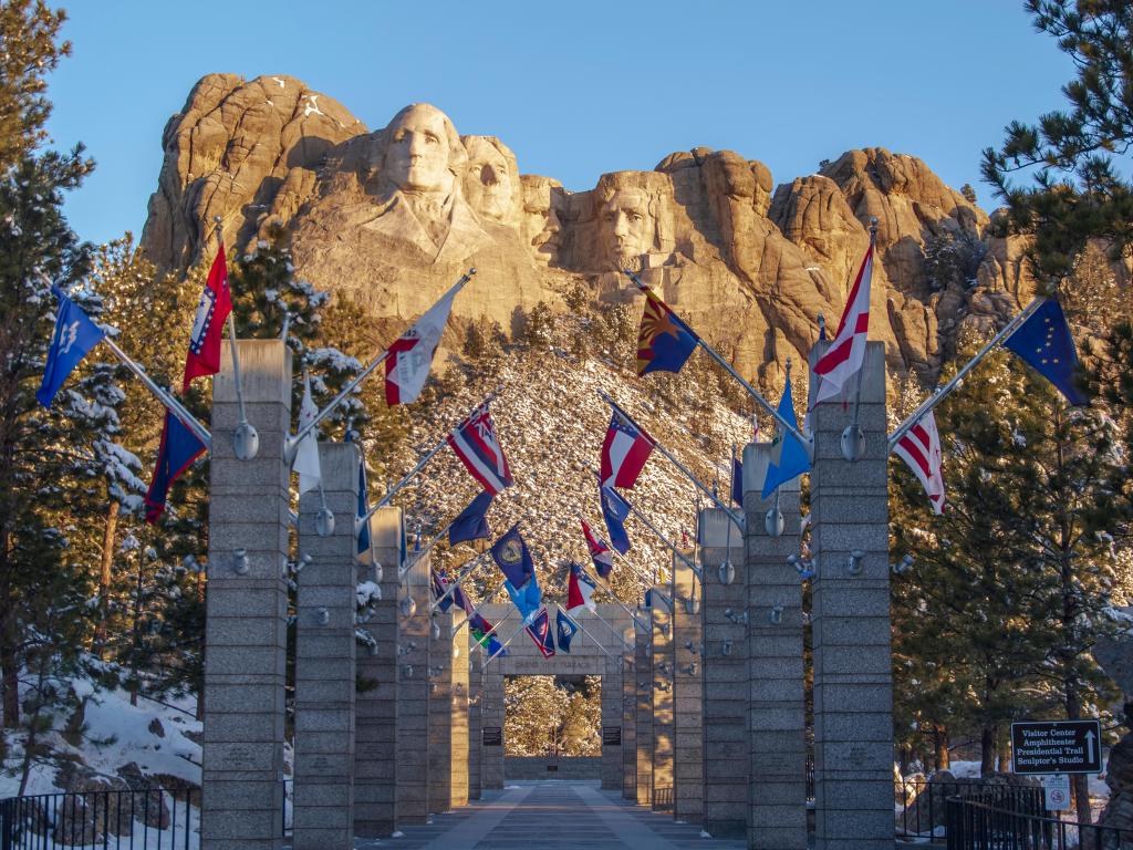 Mount Rushmore memorial in winter with snow on the ground, the flags flying with a blue sky behind