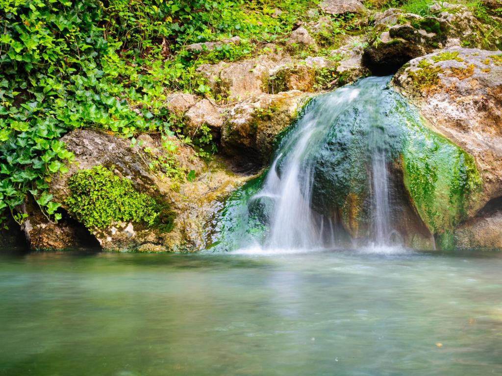 Hot Springs National Park, USA with a waterfall and rocky terrain in the background, green foliage partly covering the rocks and a river in the foreground. 