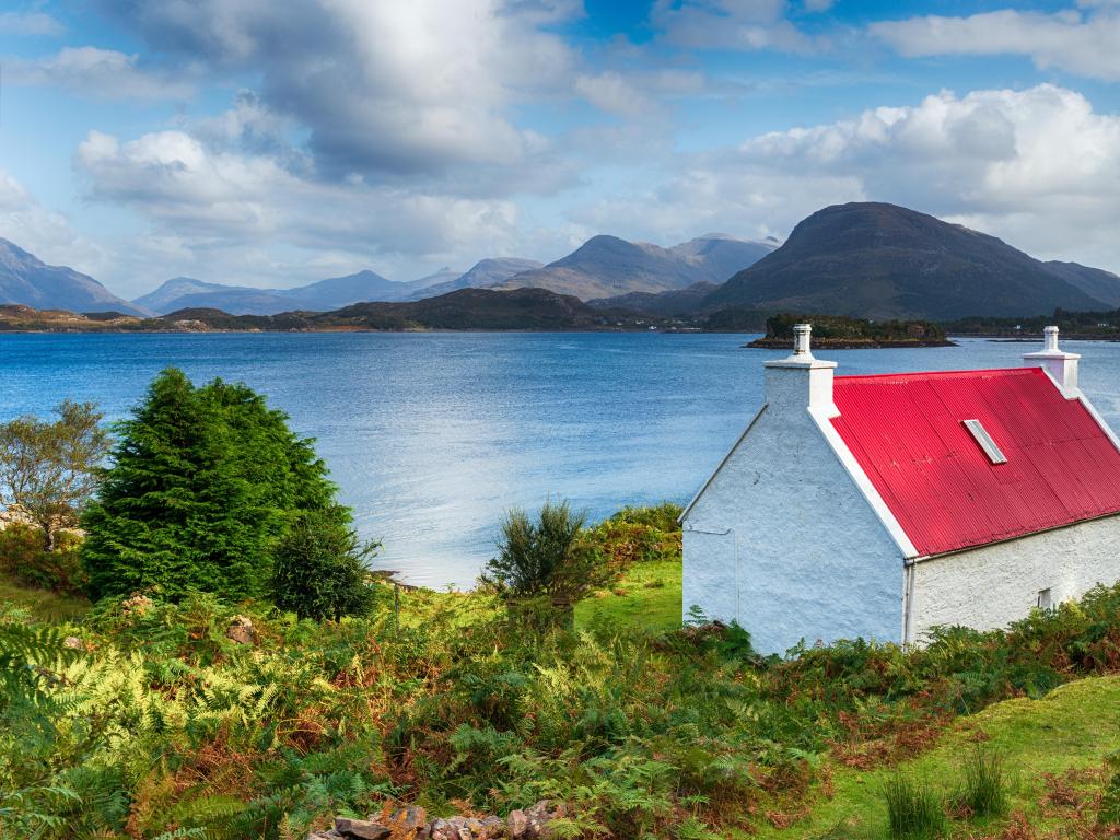 Loch Shieldaig, Highlands of Scotland, UK with a pretty red roofed croft on the shores of the loch and mountains in the distance against a blue sky.