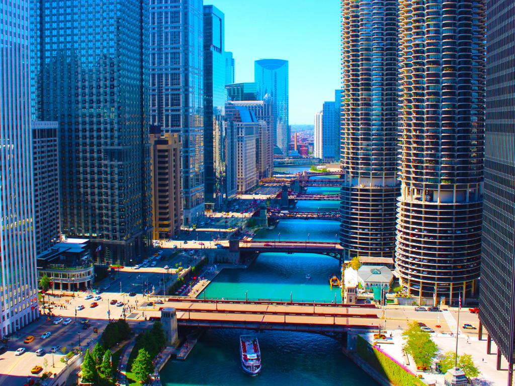 Blue river running between tall silvery blue high rise buildings with several bridges and a large road by the side of the river