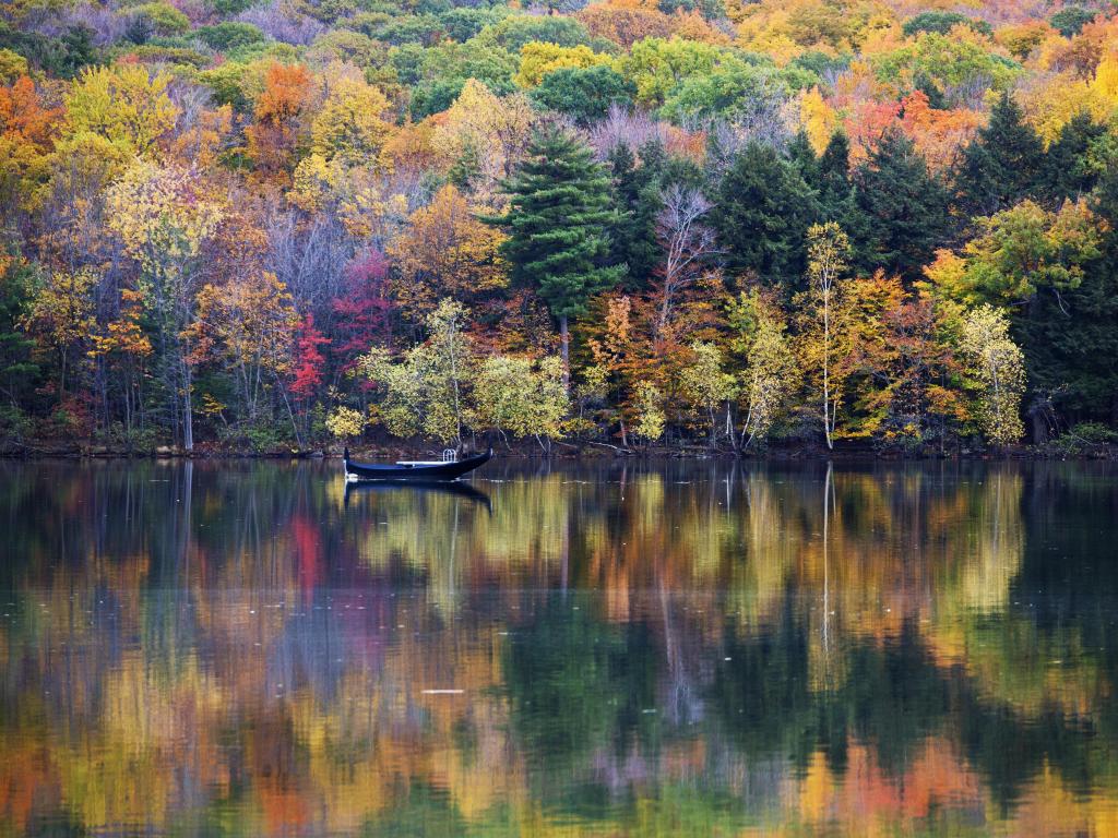Mont-Saint-Bruno National Park, Canada with fall trees in various colours reflecting in the lake in the foreground and a single boat on the water. 