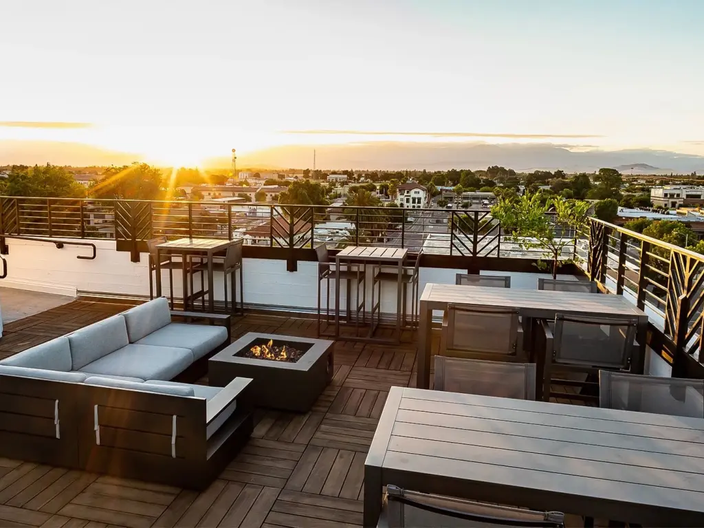 The view of the sunset from the top of a rooftop terrace with seating around a firepit and other tables and chairs 