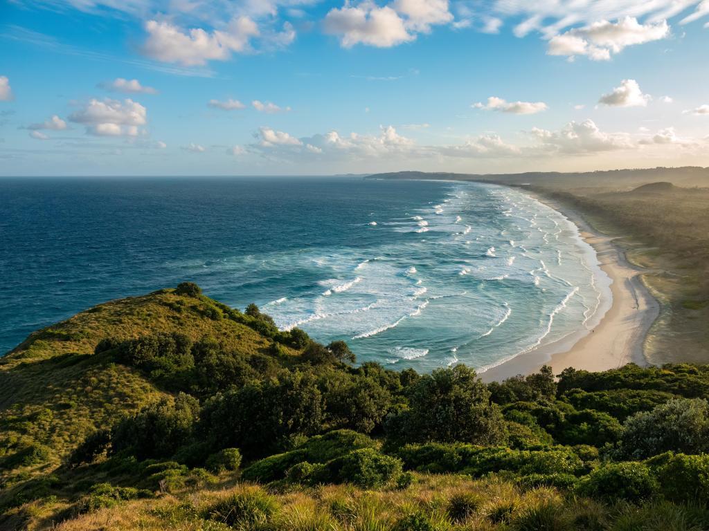 Waves crashing into the shore on a partially-cloudy day, photo taken from above a hill