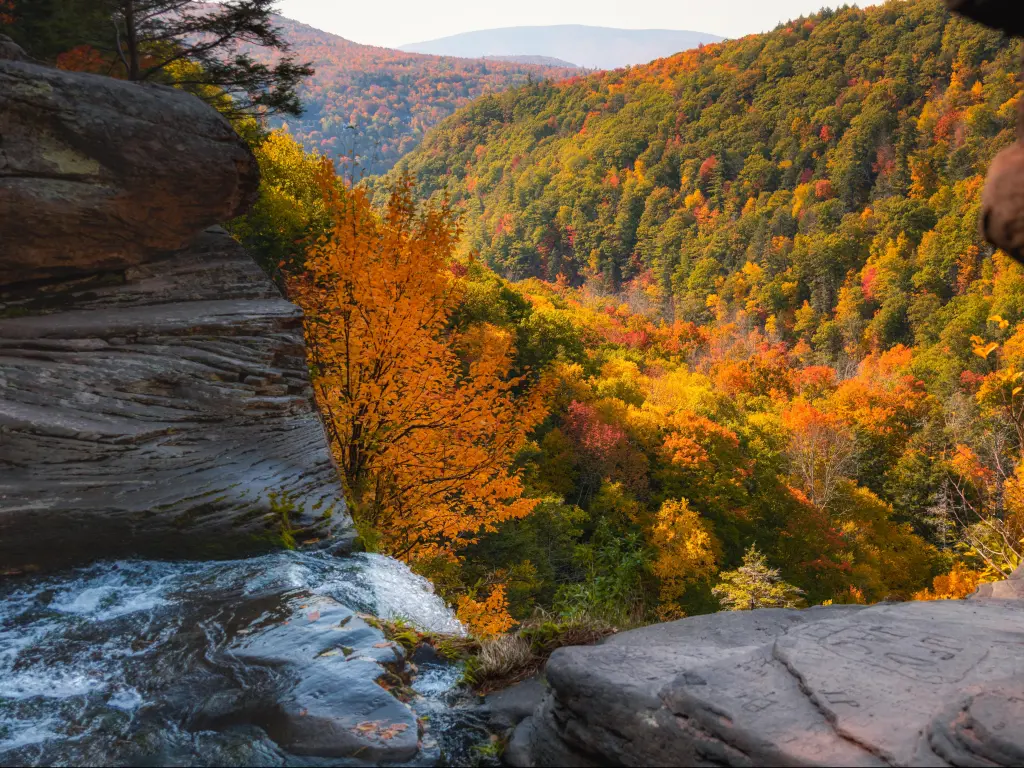 Water streaming off a rocky ledge leading into a valley valley fill with vibrant fall foliage color