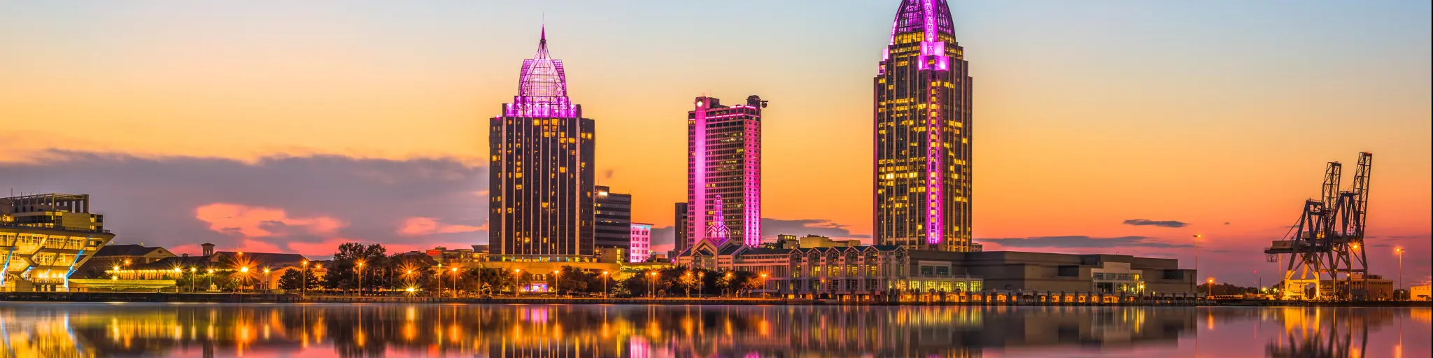 Mobile, Alabama, USA with a large expanse of water in the foreground and the downtown skyline in the distance reflecting in the water at dusk.