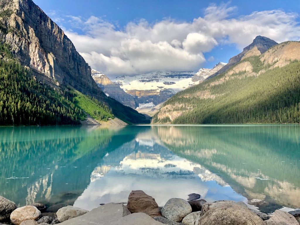 Lake Louise, Banff, Canada with rocks in the foreground, the stunning turquoise water and tree covered mountains in the distance, taken on a sunny but cloudy day.