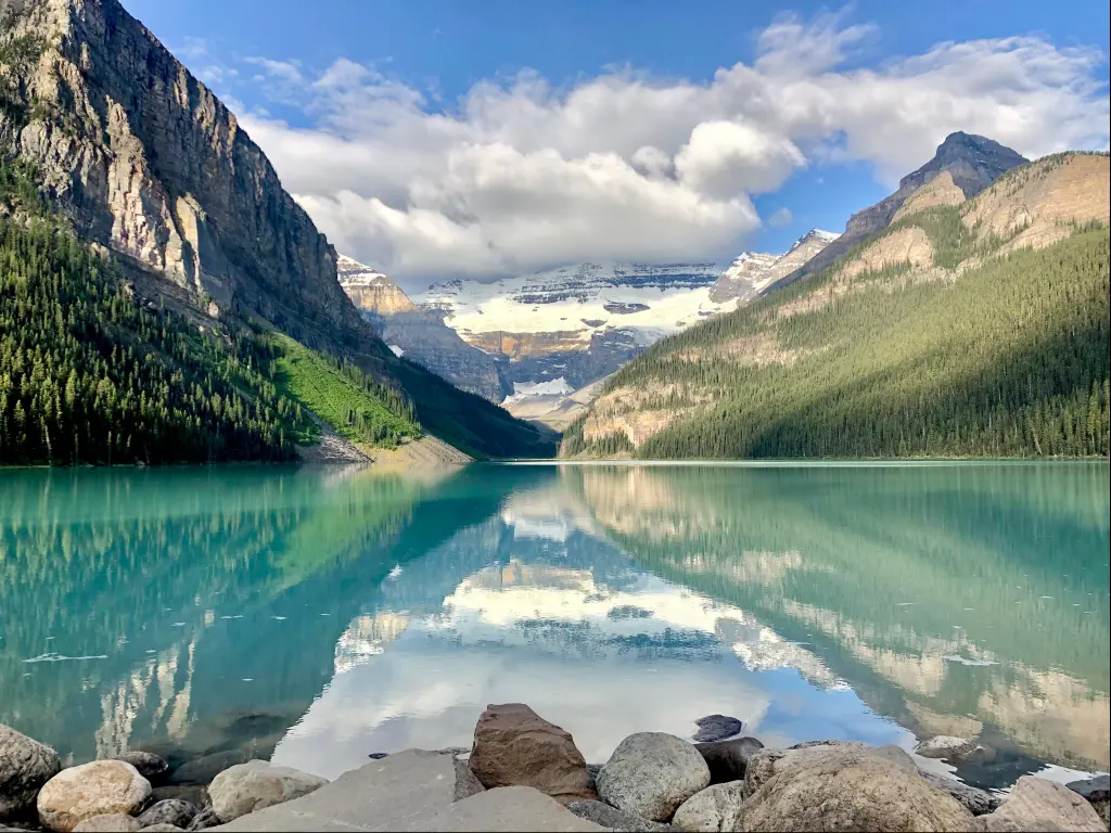 Lake Louise, Banff, Canada with rocks in the foreground, the stunning turquoise water and tree covered mountains in the distance, taken on a sunny but cloudy day.
