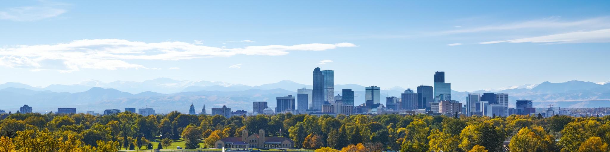 Panoramic view of high rise buildings of Denver skyline with green trees in the foreground and rocky mountains in the background