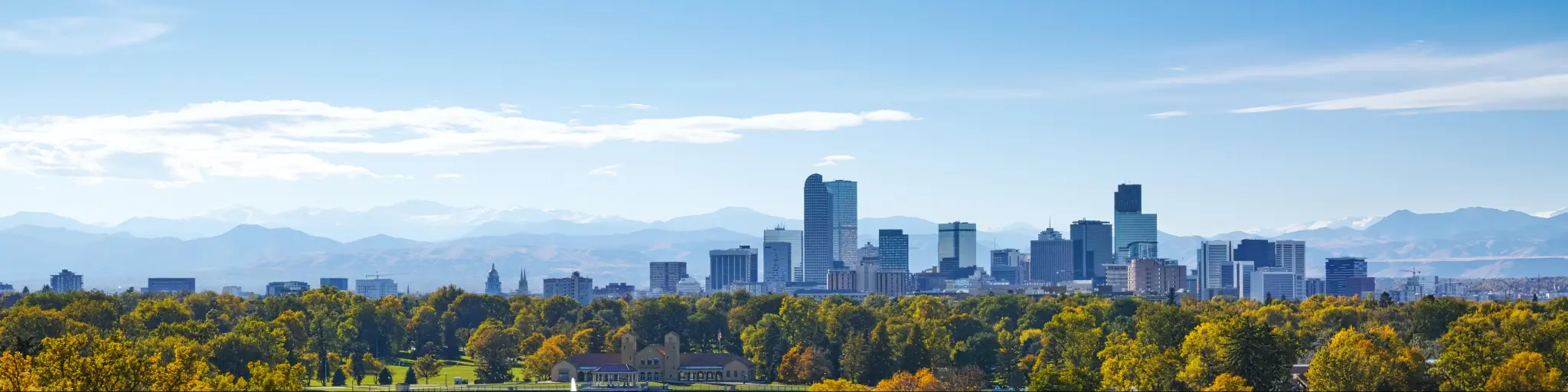 Panoramic view of high rise buildings of Denver skyline with green trees in the foreground and rocky mountains in the background