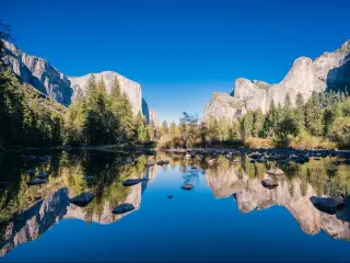 Yosemite National Park, USA on a sunny day with the lake reflecting mountains in the distance and trees lining the waters edge under a blue sky,