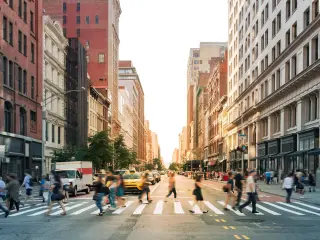 Pedestrians navigating a crosswalk at a busy intersection with the sun setting between the buildings