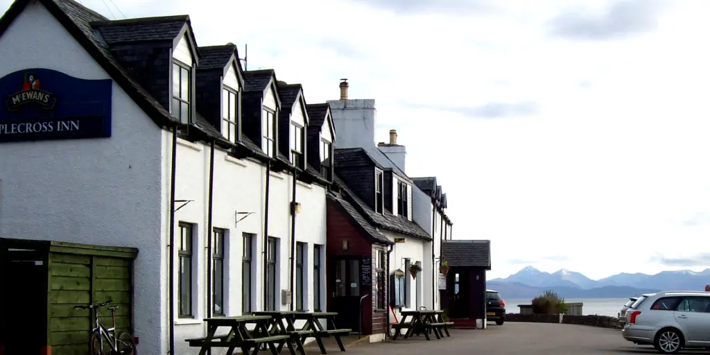 The white exterior of the Applecross in with water and the mountains in the background, and a silver car to the right