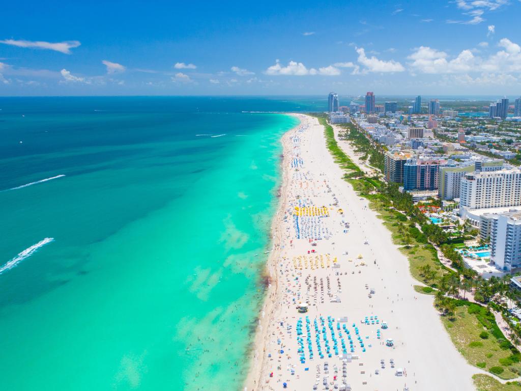 Miami Beach, Florida aerial view of the long stretch of beach with sun beds, either side of a turquoise sea and the city buildings on the right.