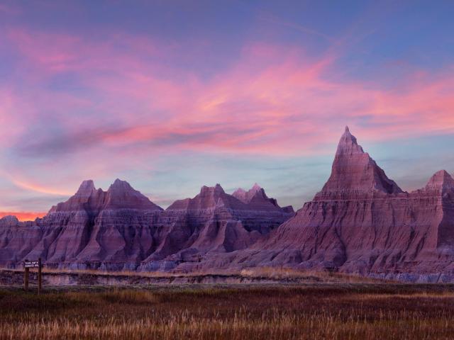 Panoramic shot showing the jagged, gray eroded peaks of Badlands National Park at sunset, with a purple-pink hued sky