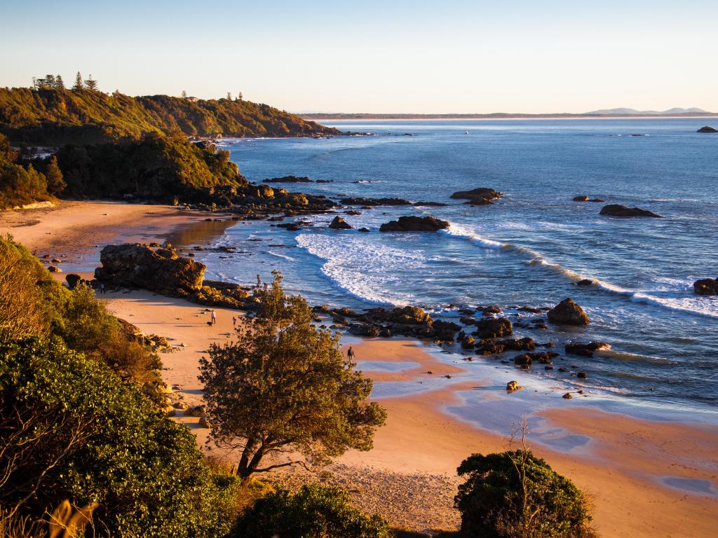 Two walkers on an otherwise empty golden sand beach with tree-covered cliffs