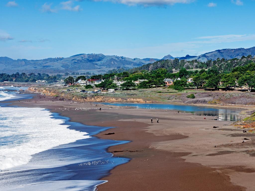 The Pacific Ocean lapping the shore at Moonstone Beach, Cambria California with blue sky above and mountains behind