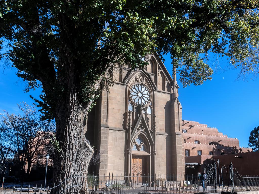 Front of Loretto Chapel in Santa Fe, with large tree in forefront on clear, blue sky day.