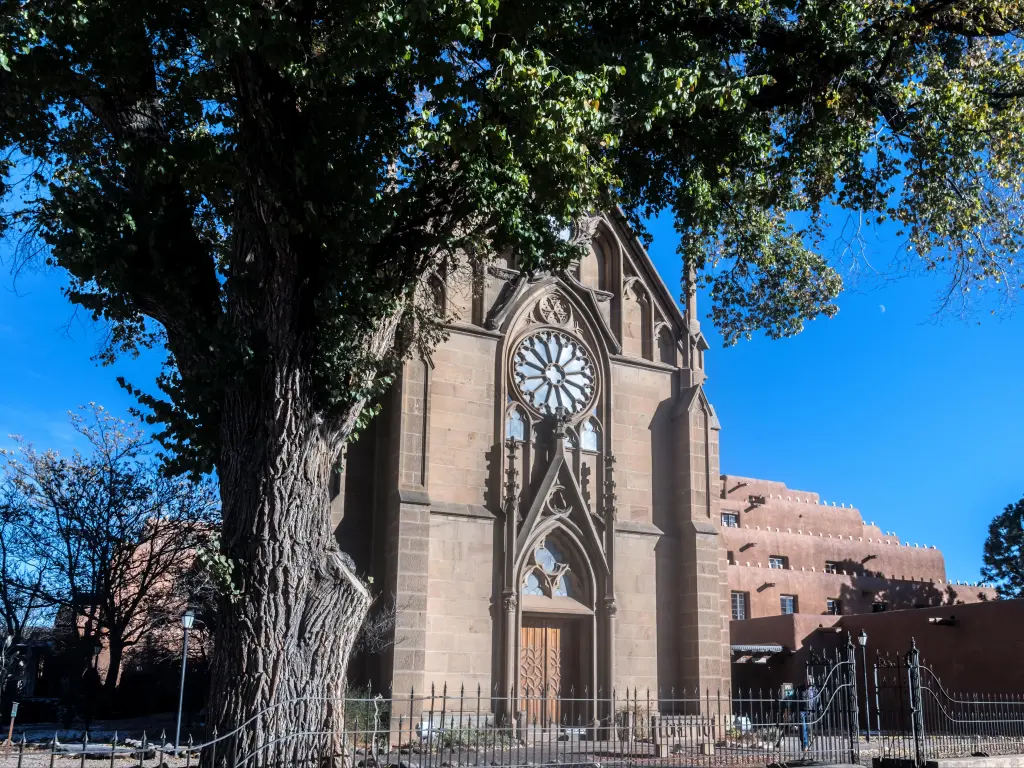 Front of Loretto Chapel in Santa Fe, with large tree in forefront on clear, blue sky day.