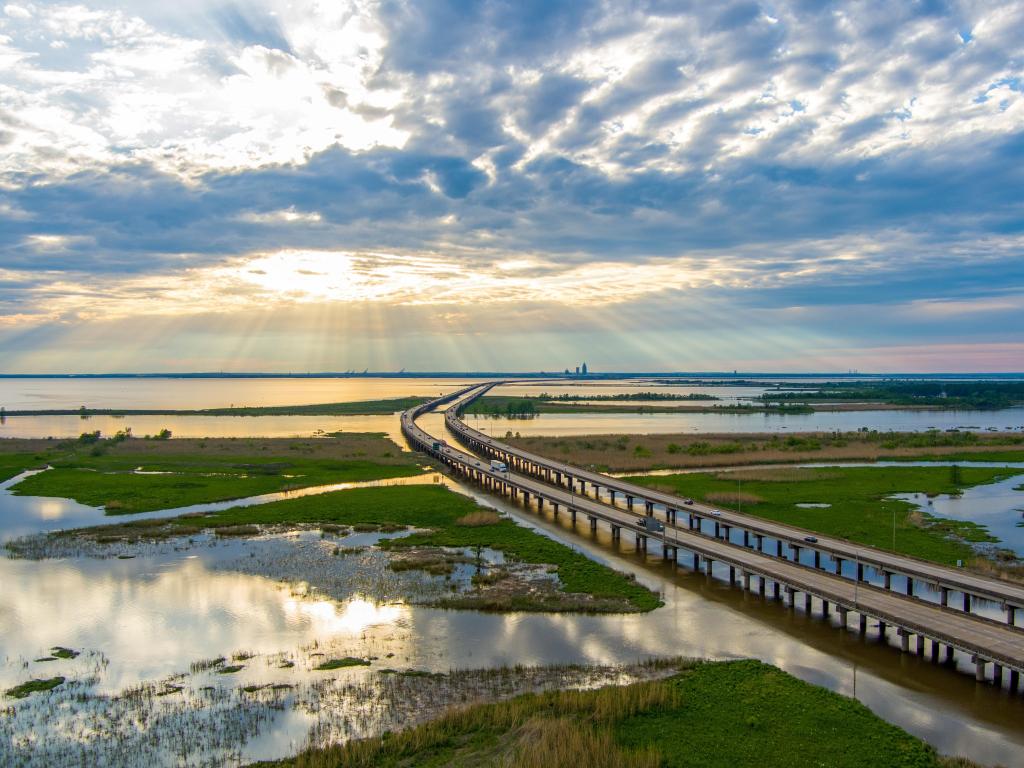 Mobile Bay, Alabama, USA taken as an aerial view of the bay at sunset, with grassy and water surrounding the bridge leading into the distance.