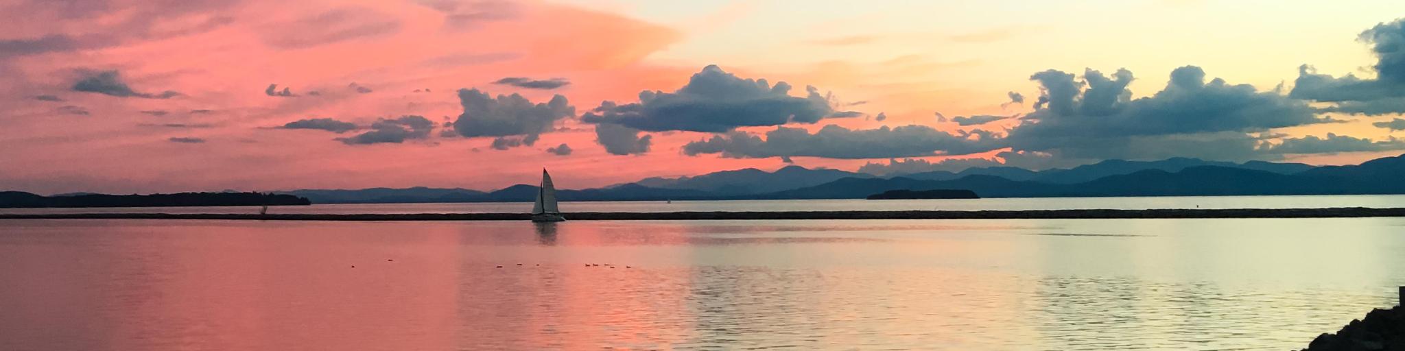 Lake Champlain in Burlington, Vermont, USA with a sailboat at sunset on Lake Champlain in Burlington, Vermont. The vibrant pink sky transitions to a cool blue in both the sky and the water.