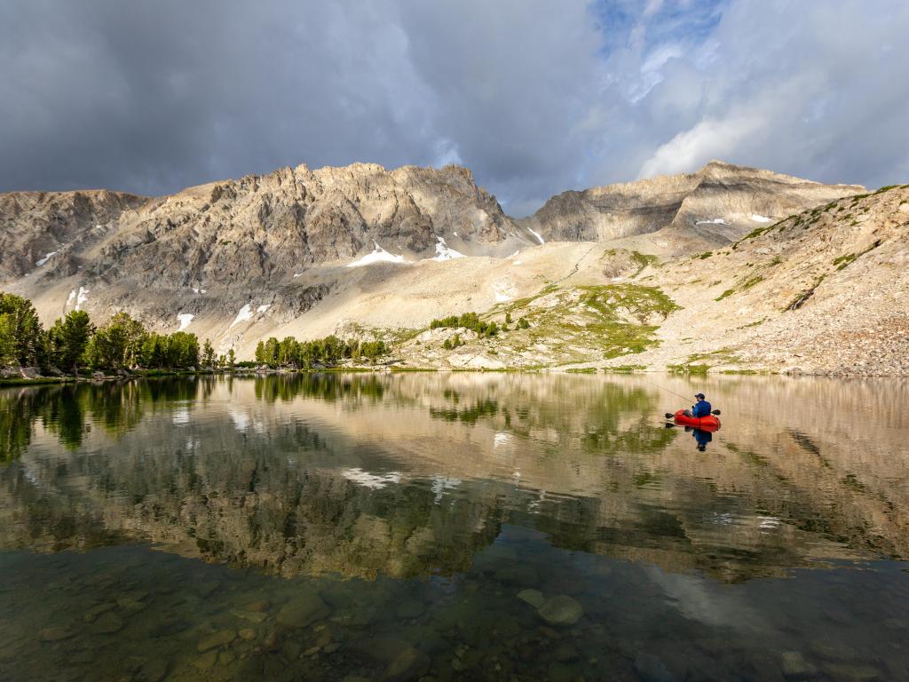Sun Valley, Idaho, USA with someone fly fishing from a pack raft in an alpine lake in the foreground, mountains and clouds above.