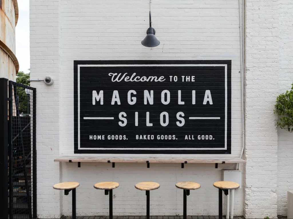 Famous Magnolia Silos in Waco, a seating area with stools facing a white brick wall with a black sign that says "Magnolia Silos"