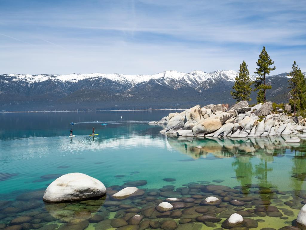 Sand Harbor Beach, Lake Tahoe, USA with small and large rocks in the foreground surrounded by turquoise water, snow-capped mountains in the distance on a sunny day with a few paddle boards in the water.