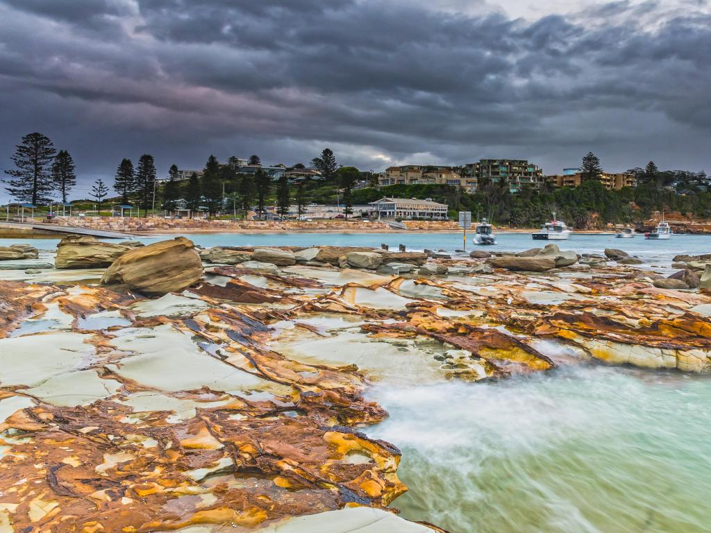 Terrigal, NSW, Australia with rocks and sea in the foreground, and buildings and trees in the background below a cloudy sky.