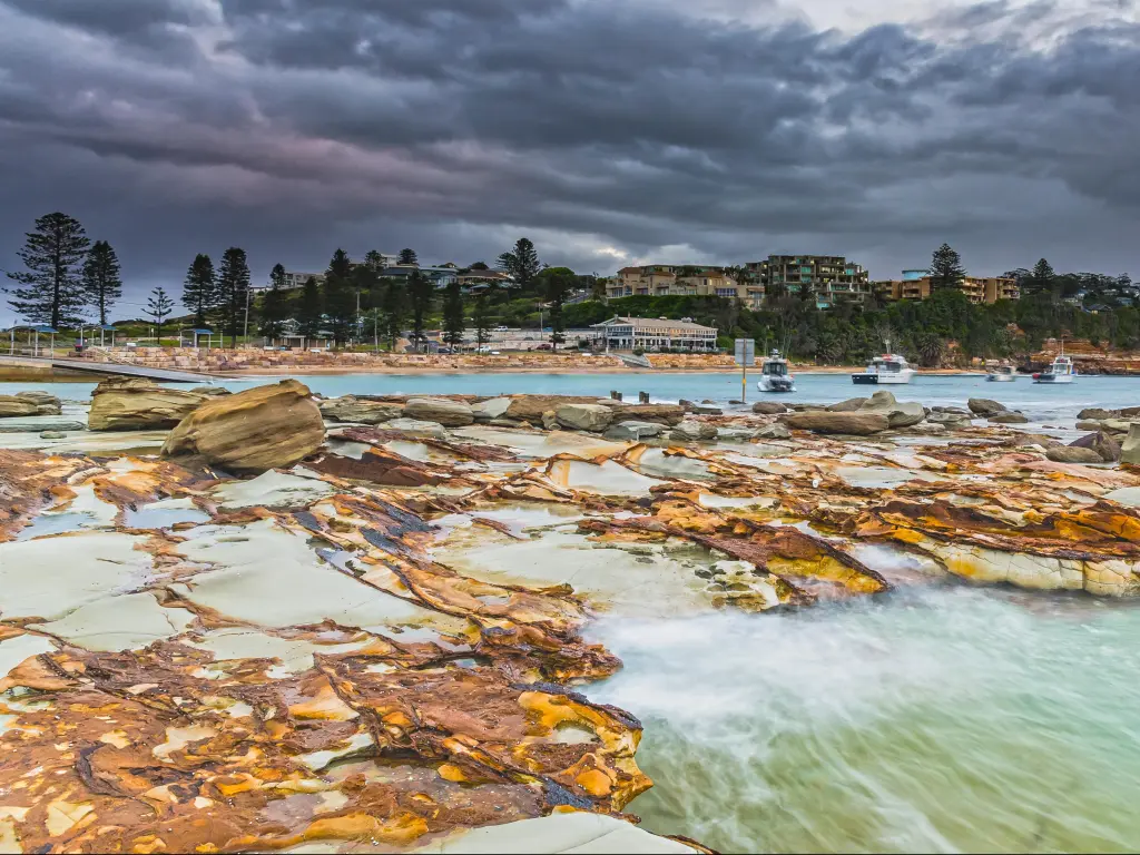 Terrigal, NSW, Australia with rocks and sea in the foreground, and buildings and trees in the background below a cloudy sky.