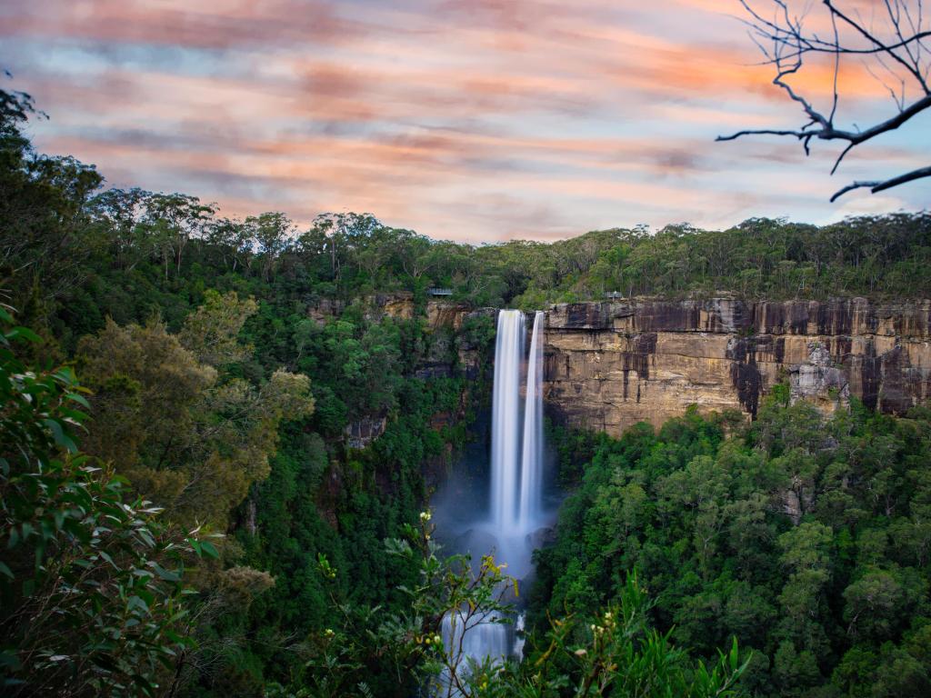 High waterfalls over sheer cliff edge with exposed rock face and trees under an orange and pink sky