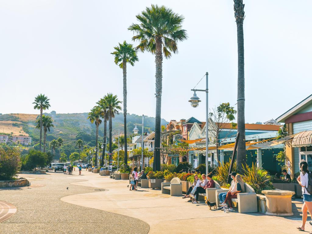 Beachfront of Avila Beach, with people sitting outside various colorful fronted restaurants and shops along the palm-tree lined promenade
