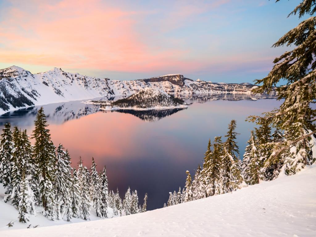 Crater Lake, Oregon, with a pink hued sunset in winter, with snow capped mountains in the background and trees around the edge
