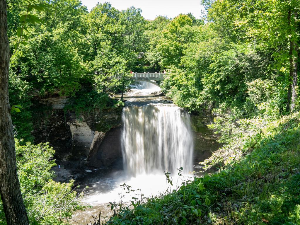 Minneopa Falls, Minneopa State Park, Mankato, Minnesota with a waterfall central in the image and tall trees and greenery all around and a white wooden bridge in the distance. 